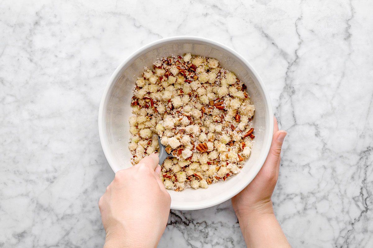 overhead shot of a person’s hands mix a crumbly mixture of flour, sugar, and pecans in a large white bowl on a marble countertop