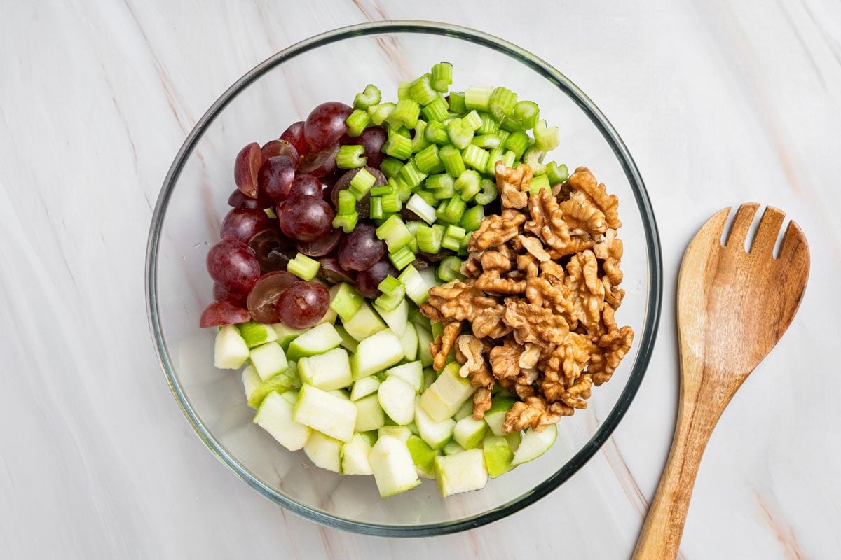 Overhead shot of combine the apples, grapes, celery and walnuts in a large glass bowl; a wood spoon resting on a marble surface;