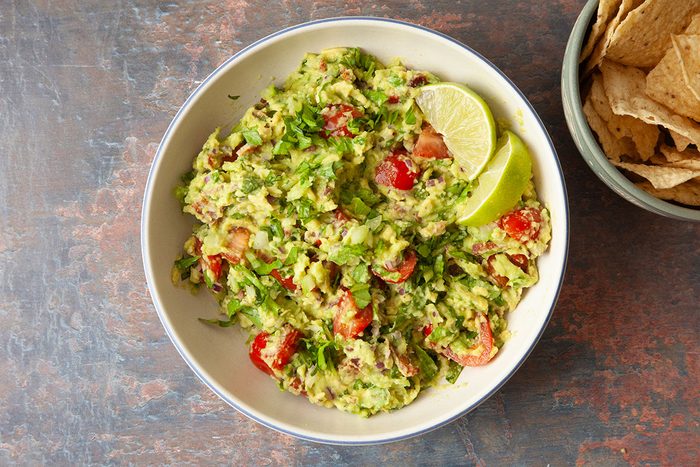 A bowl of guacamole with chopped tomatoes, onions, cilantro, and two lime wedges, next to a bowl of tortilla chips on a rustic surface.