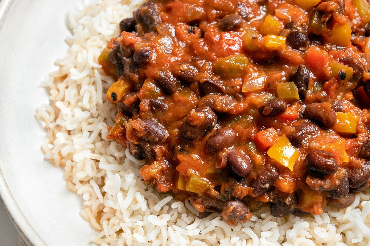 A close-up of a plate with cooked brown or white rice topped with a chunky vegetable and black bean stew in a rich tomato-based sauce.