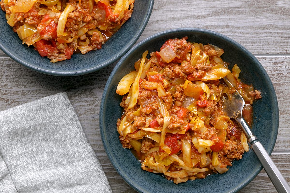 Two blue bowls filled with a mixture of cooked cabbage, ground meat, tomatoes, and onions sit on a wooden surface next to a folded gray napkin. A fork rests in one of the bowls.