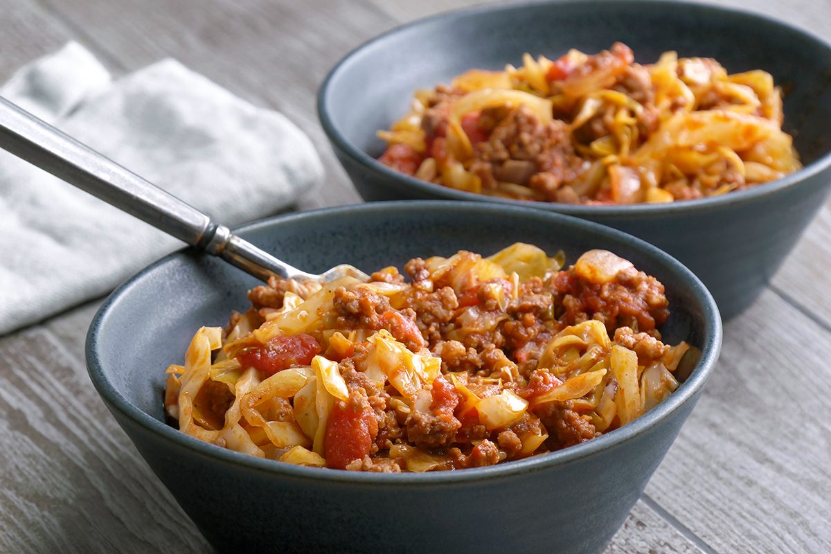 Two dark bowls filled with pasta topped with a chunky meat and tomato sauce, sitting on a wooden table with a folded white napkin nearby. One bowl has a fork resting inside.