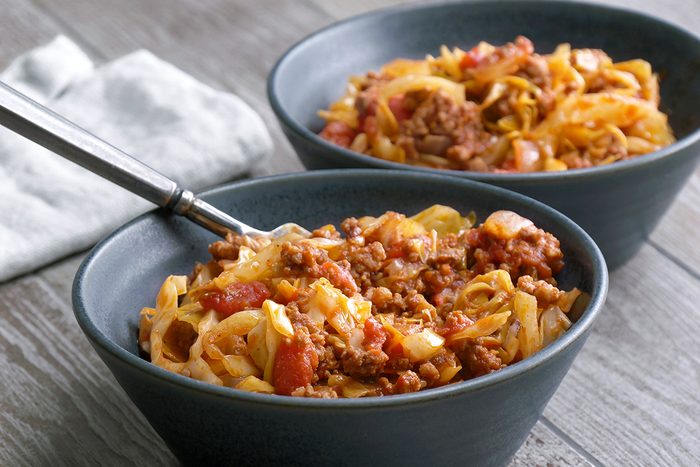 Two dark bowls filled with pasta topped with a chunky meat and tomato sauce, sitting on a wooden table with a folded white napkin nearby. One bowl has a fork resting inside.