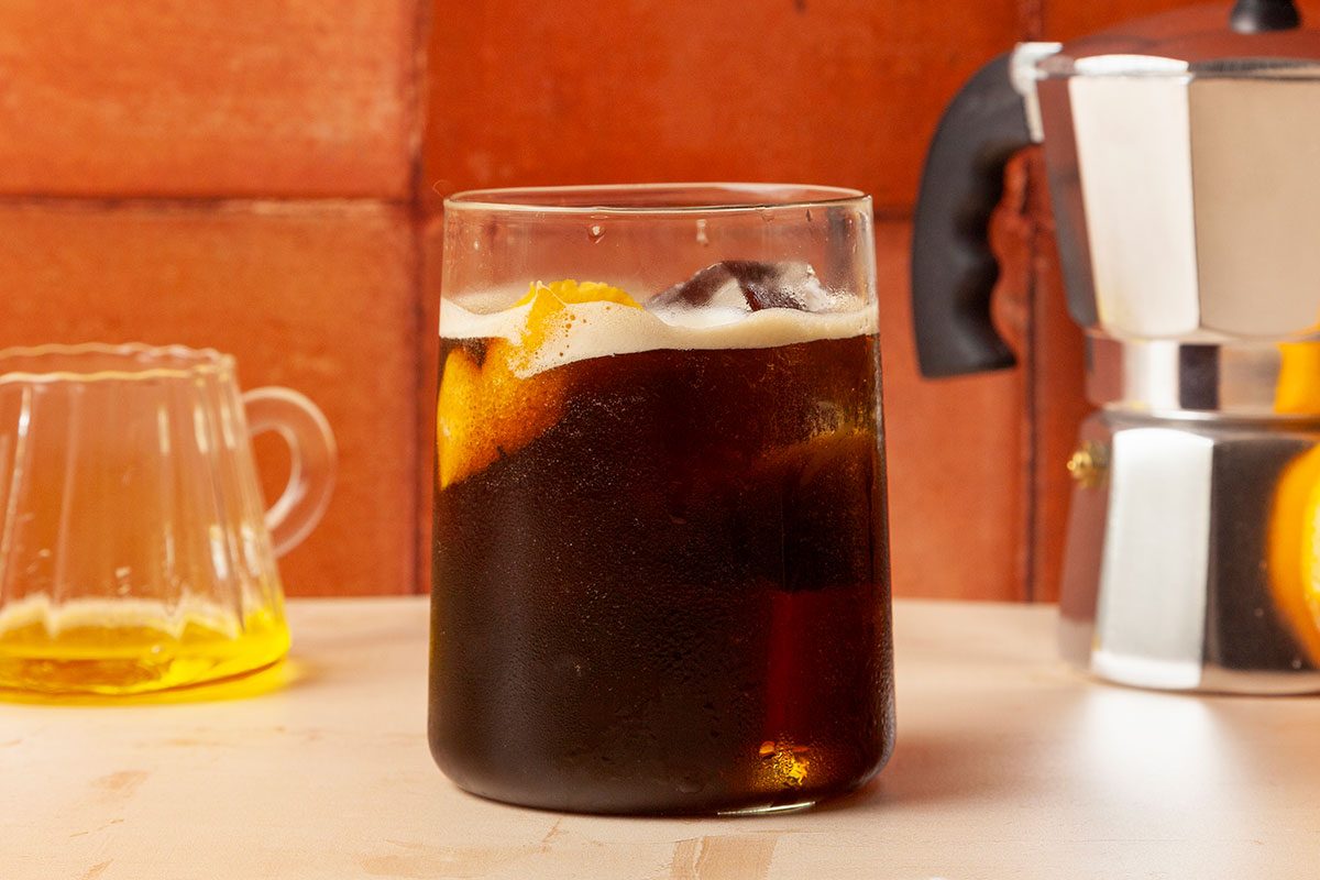 Table view shot of Carajillo filled in a glass; a small glass pitcher containing liquor and cocktail shaker are visible nearby; all set on a painted surface;