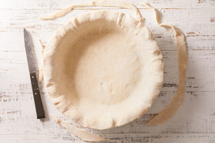 A round pie dish lined with unbaked pie crust sits on a white wooden surface. A knife and trimmed excess dough are next to the dish.