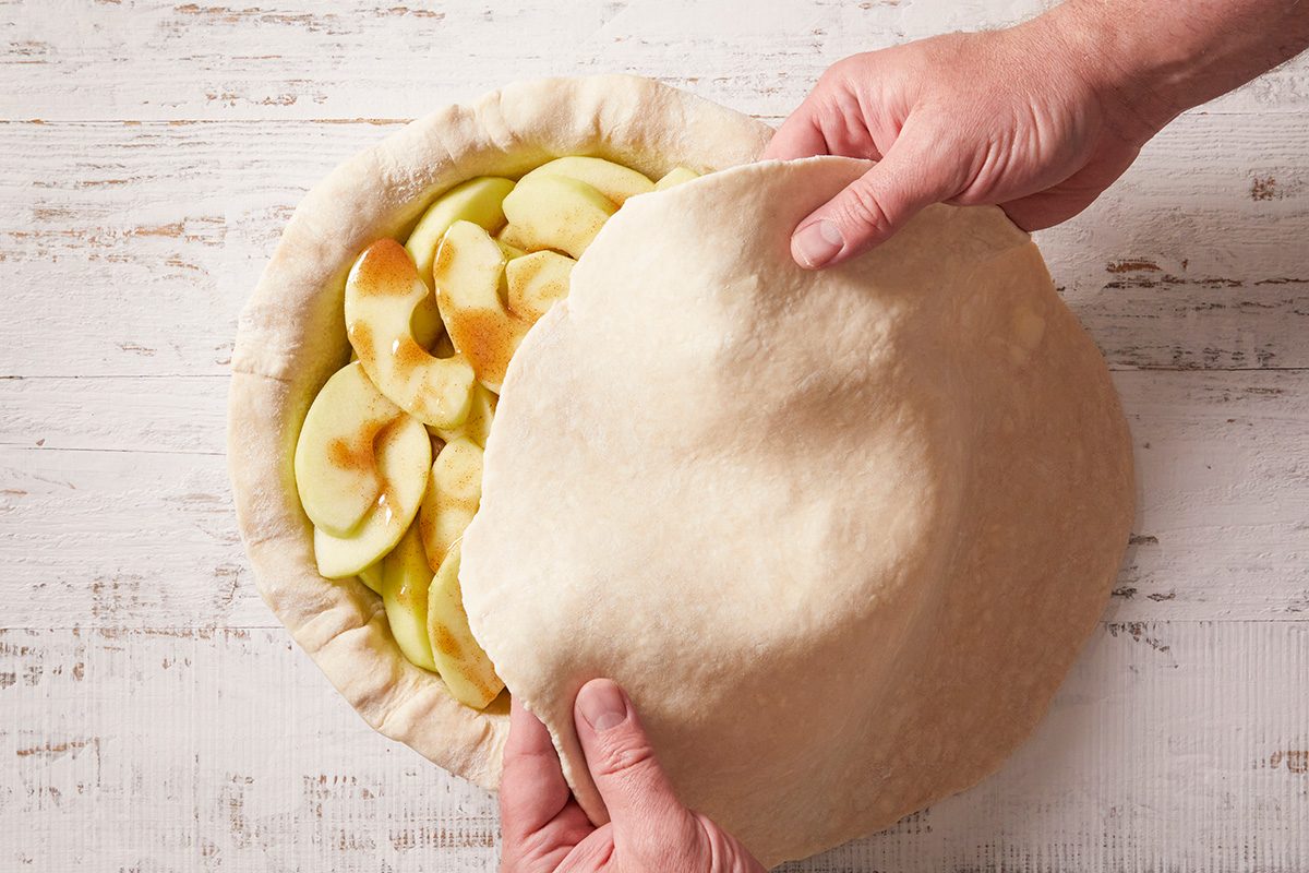 A person places a sheet of pie dough over sliced apples in a pie crust on a white wooden surface, preparing to cover the filling before baking.