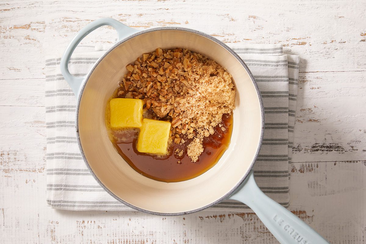 A white enamel pot with a blue handle contains brown sugar, two cubes of butter, and liquid syrup, sitting on a striped cloth over a white wooden surface.