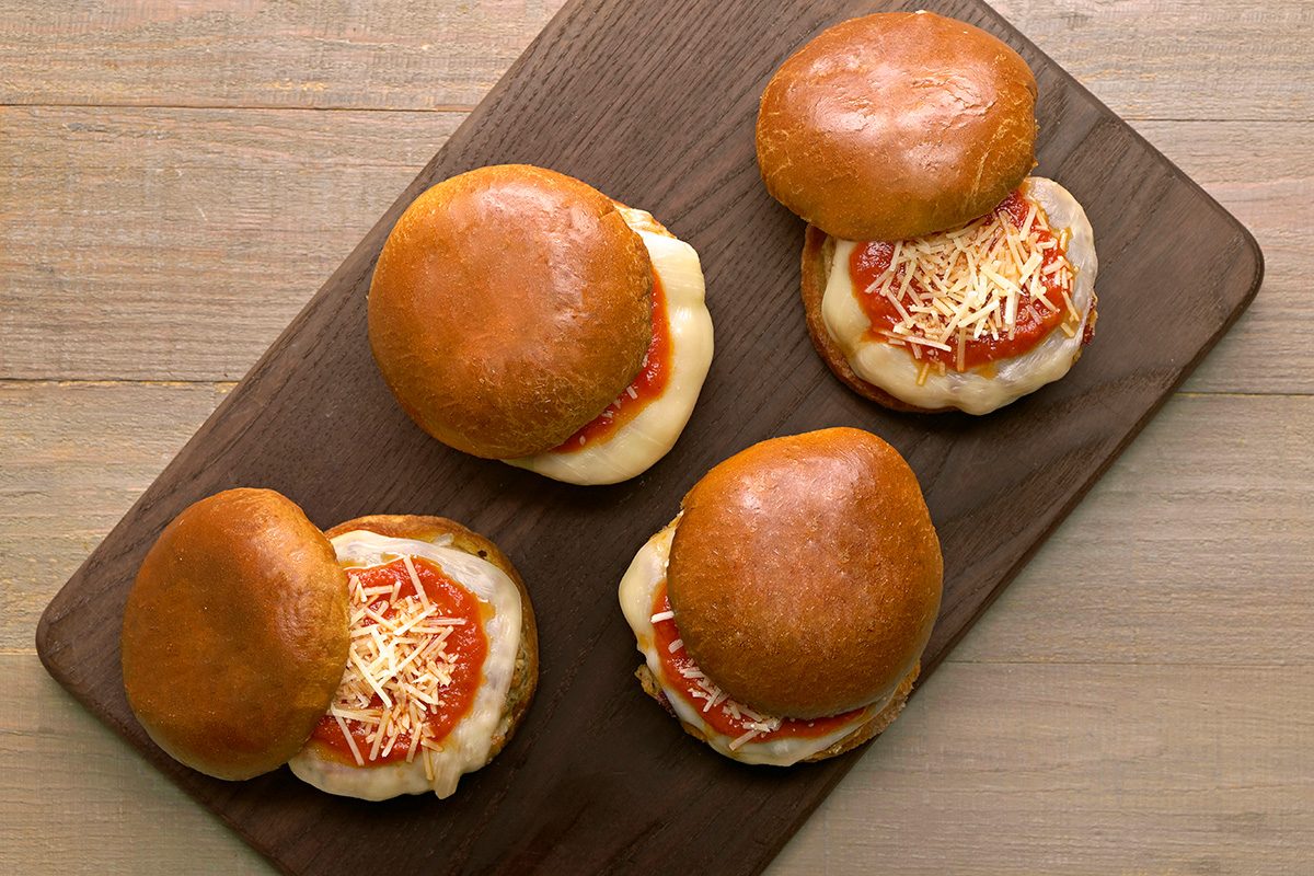 Four sandwiches with golden buns, melted cheese, tomato sauce, and shredded cheese are arranged on a dark wooden board, viewed from above on a light wood surface.