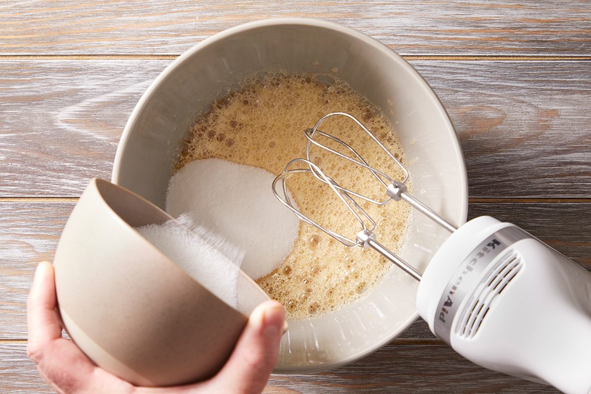 A hand pours sugar from a bowl into a mixing bowl with a frothy liquid, while holding an electric hand mixer above the bowl on a wooden surface.