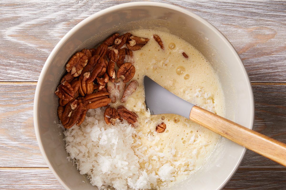 A mixing bowl with shredded coconut, pecan halves, and a creamy liquid mixture being stirred with a spatula on a wooden surface.