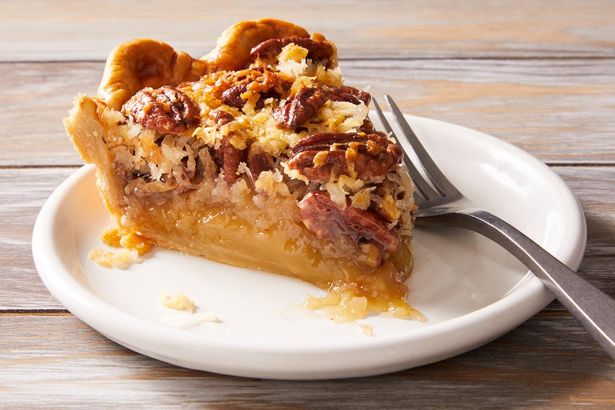 A slice of pecan pie with a golden, flaky crust and a gooey filling sits on a white plate with a fork beside it, atop a wooden table.