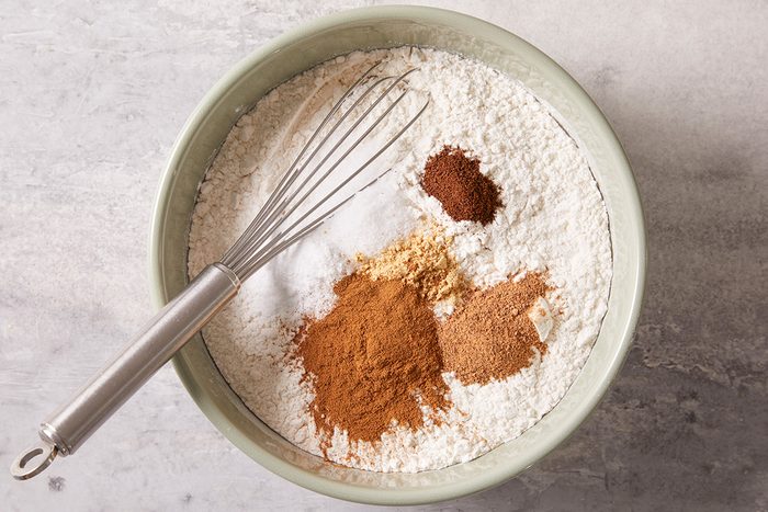 A mixing bowl with flour, baking soda, and piles of ground spices, including cinnamon and ginger, sits on a countertop. A metal whisk rests inside the bowl, ready for mixing.