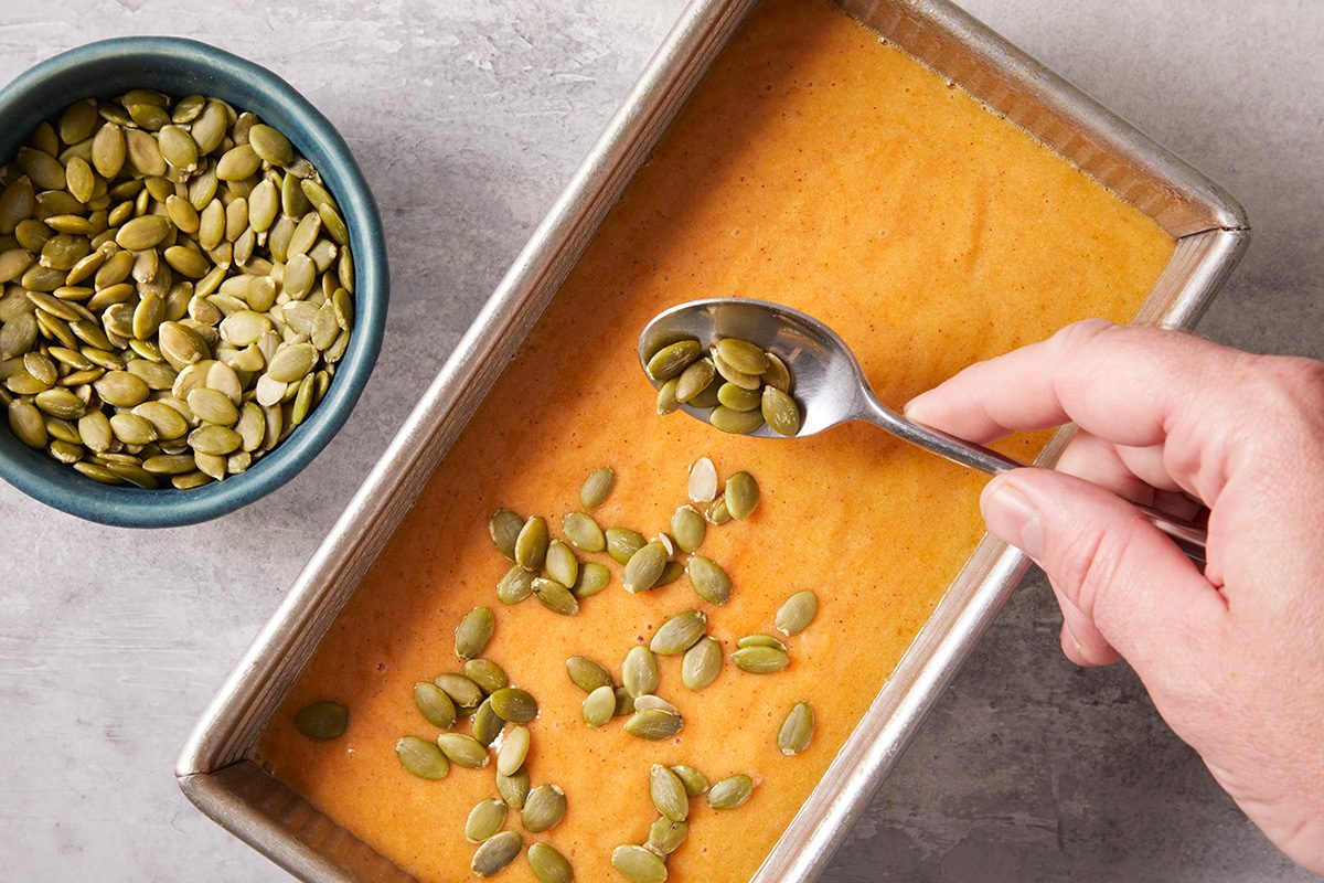 A hand sprinkles pumpkin seeds from a spoon onto an orange batter in a rectangular baking pan. A small bowl of pumpkin seeds sits nearby on a gray surface.