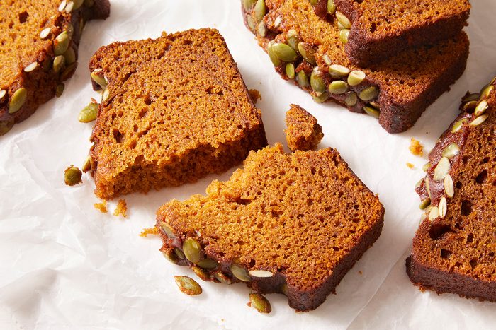 Sliced pumpkin bread with a browned crust and seeds on top, arranged on white parchment paper. One slice is broken into pieces, showing the moist texture inside.