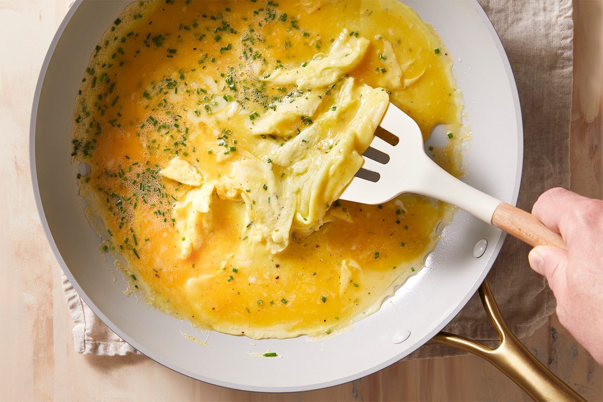 A hand uses a white spatula to gently scramble eggs in a light-colored nonstick pan. The eggs are partially cooked and sprinkled with chopped herbs. The pan sits on a cloth on a wooden surface.