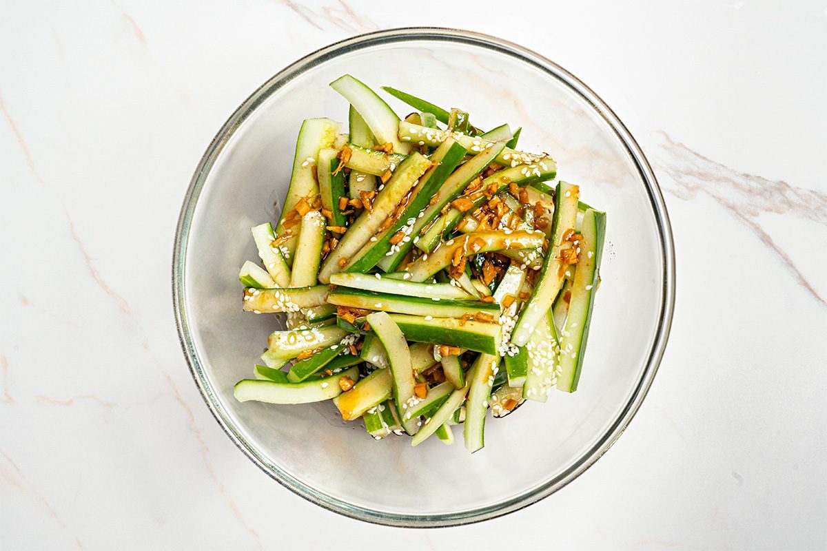 A glass bowl filled with cucumber sticks topped with a soy-based dressing, chopped garlic, and sesame seeds, placed on a white marble surface.