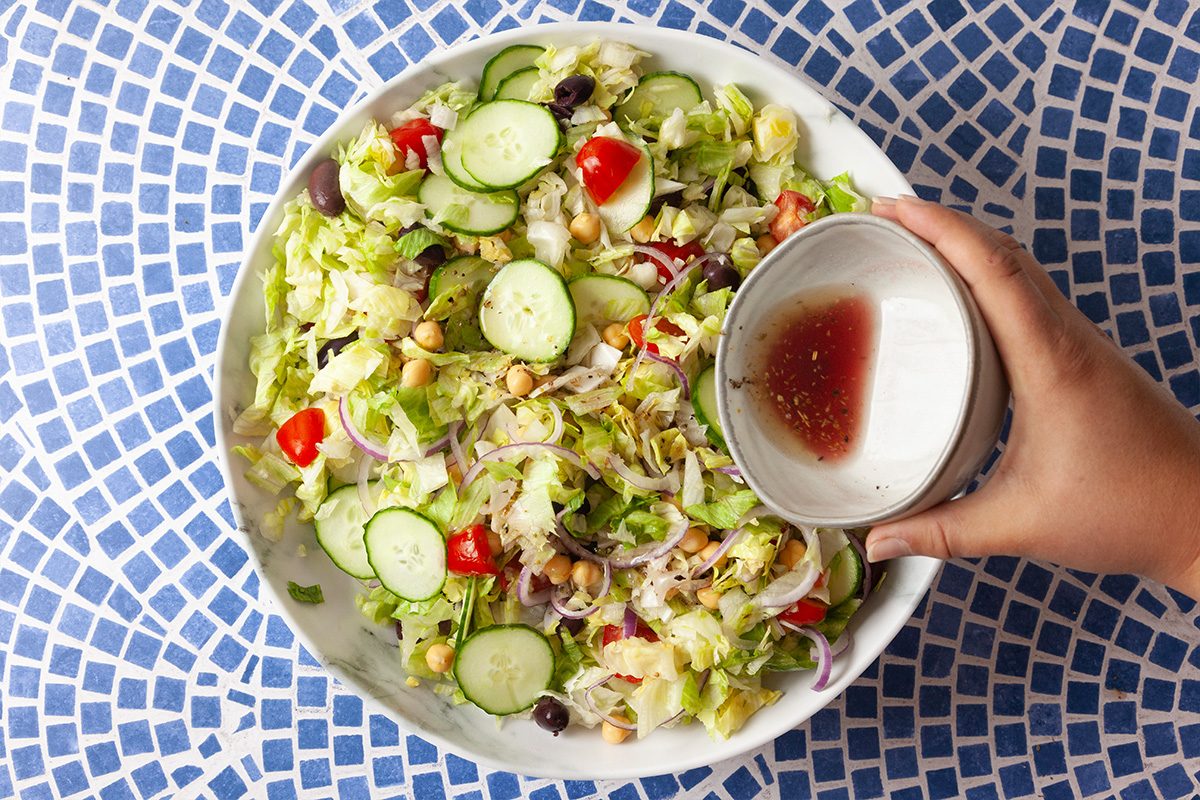 A hand pours dressing onto a fresh salad with cucumbers, tomatoes, onions, olives, and lettuce in a white bowl, placed on a blue and white patterned surface.