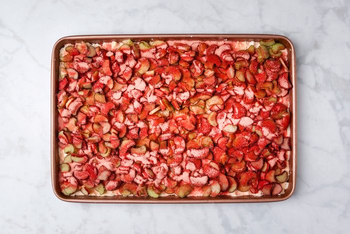 overhead shot of a baking sheet filled with an uncooked dessert topped with chopped strawberries and rhubarb, set on a light marble surface
