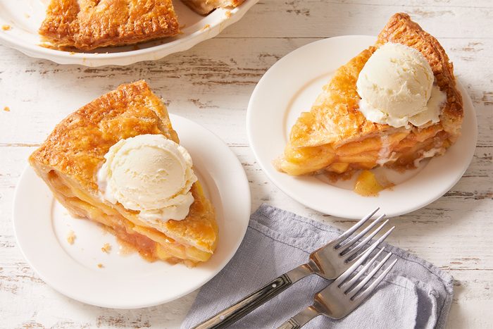Two slices of apple pie topped with vanilla ice cream are served on white plates, accompanied by two forks on a folded blue napkin on a rustic white wooden table.