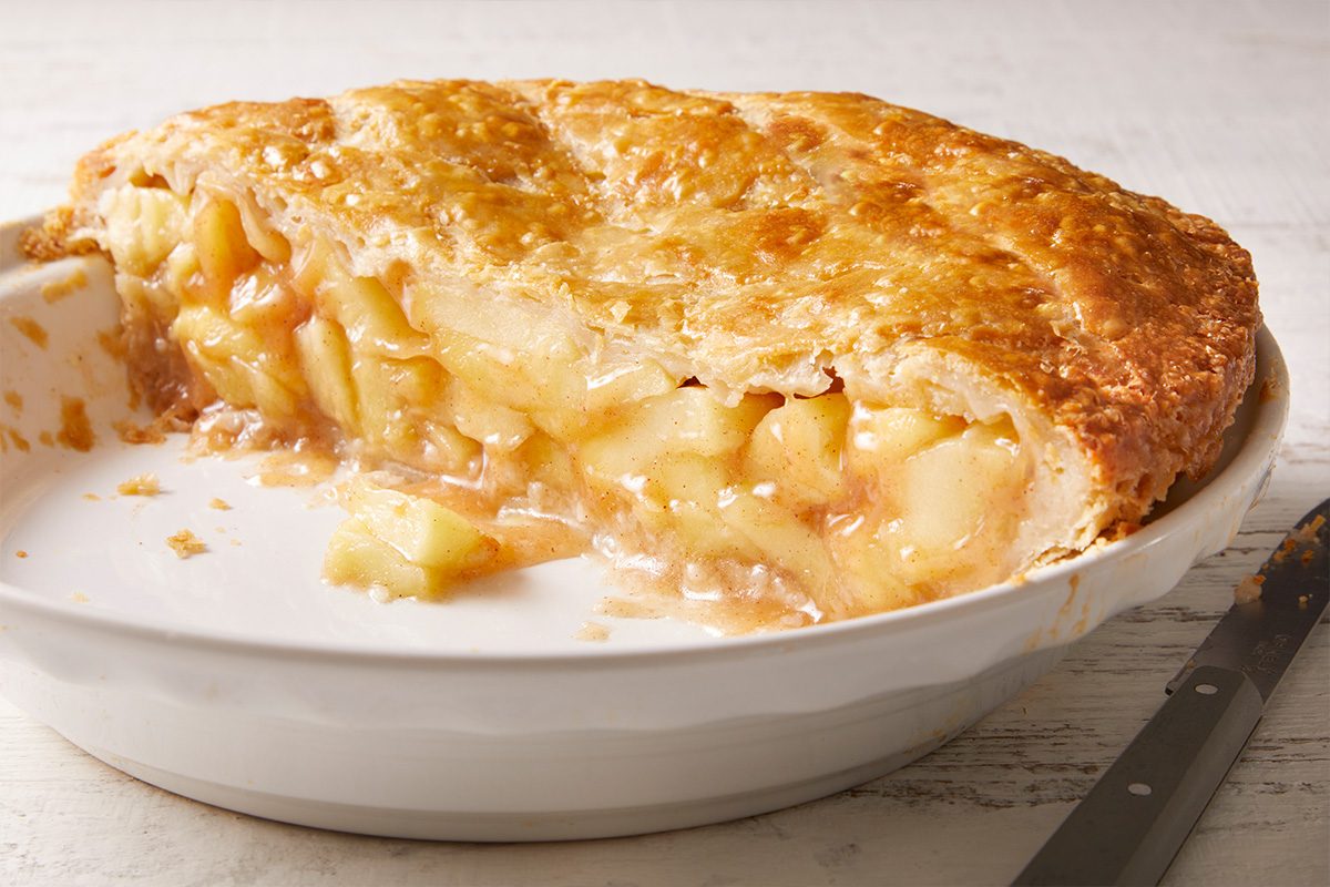 A golden-brown apple pie with a flaky crust sits in a white pie dish. A large slice has been removed, revealing the juicy, baked apple filling inside. A pie server rests nearby.