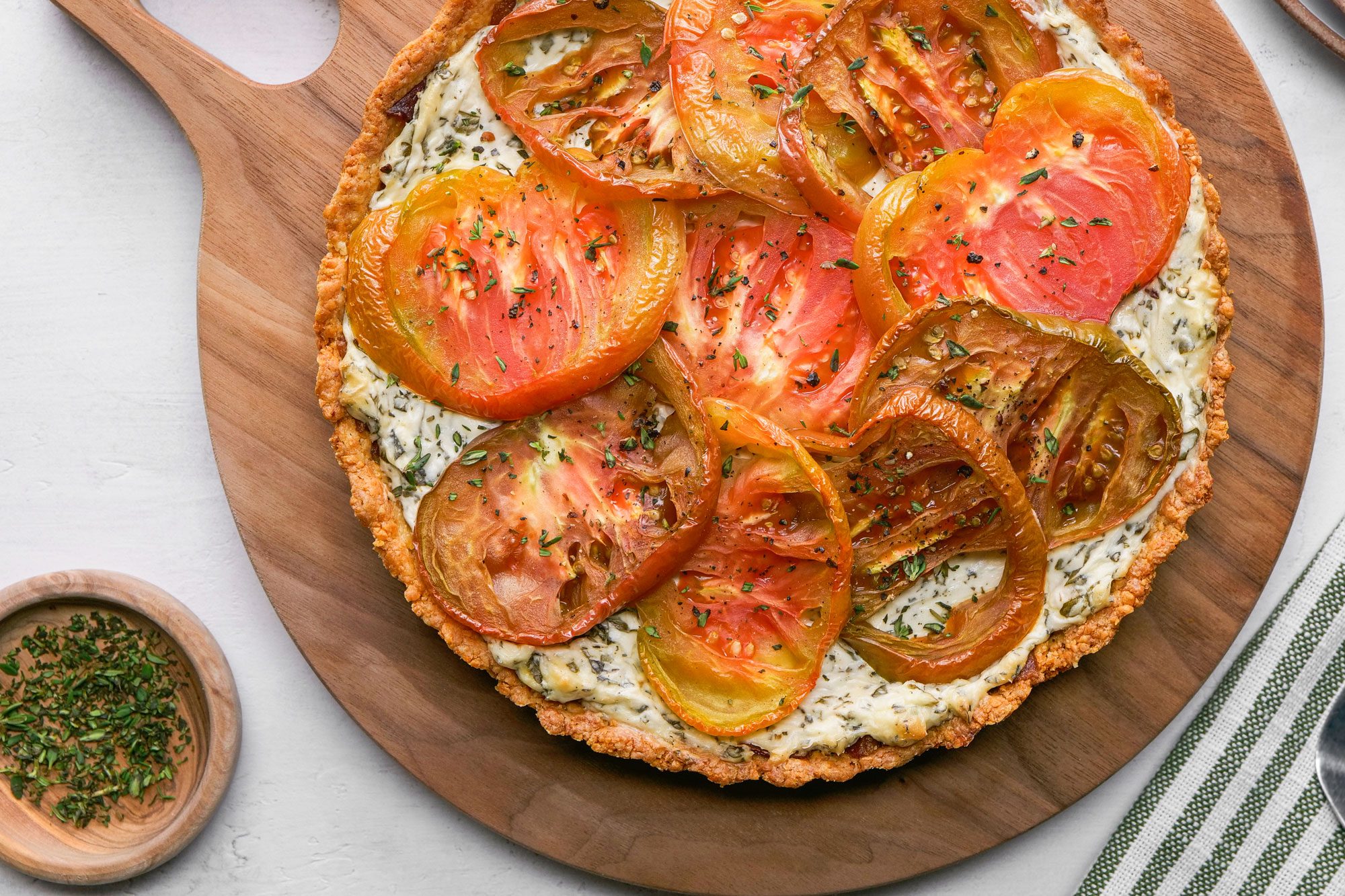 A top shot of a vibrant tomato Pie with herbed cheese filling rests on a wooden board; heirloom tomatoes decorate the top; nearby are stacked plates; a striped napkin; and a bowl of fresh herbs, creating an inviting rustic presentation