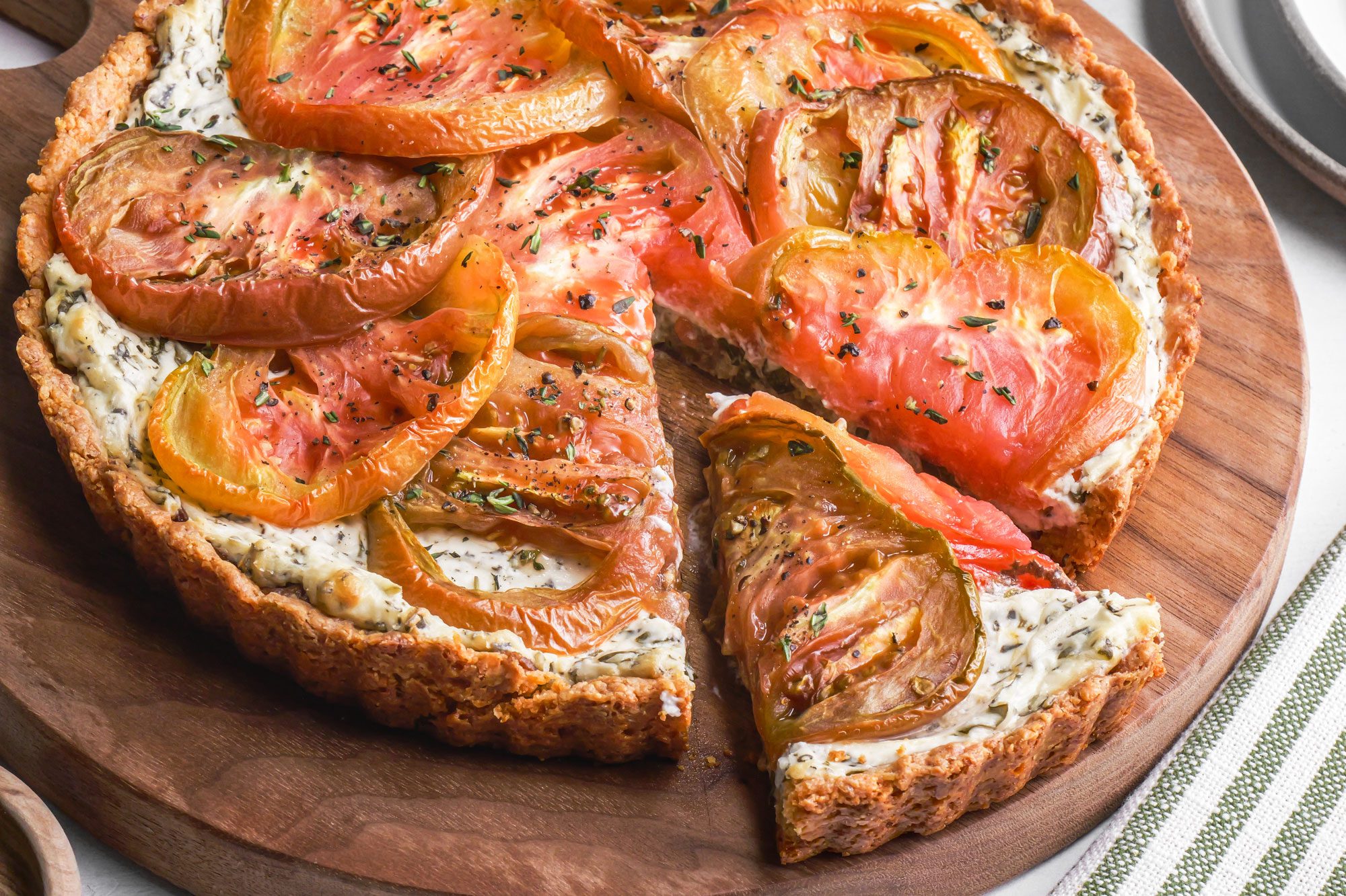 A 3/4th Shot of golden-brown Tomato pie with sliced heirloom tomatoes and fresh herbs rests on a wooden board; one slice is pulled out revealing creamy herbed filling; a striped napkin is beside it, creating a rustic and inviting scene