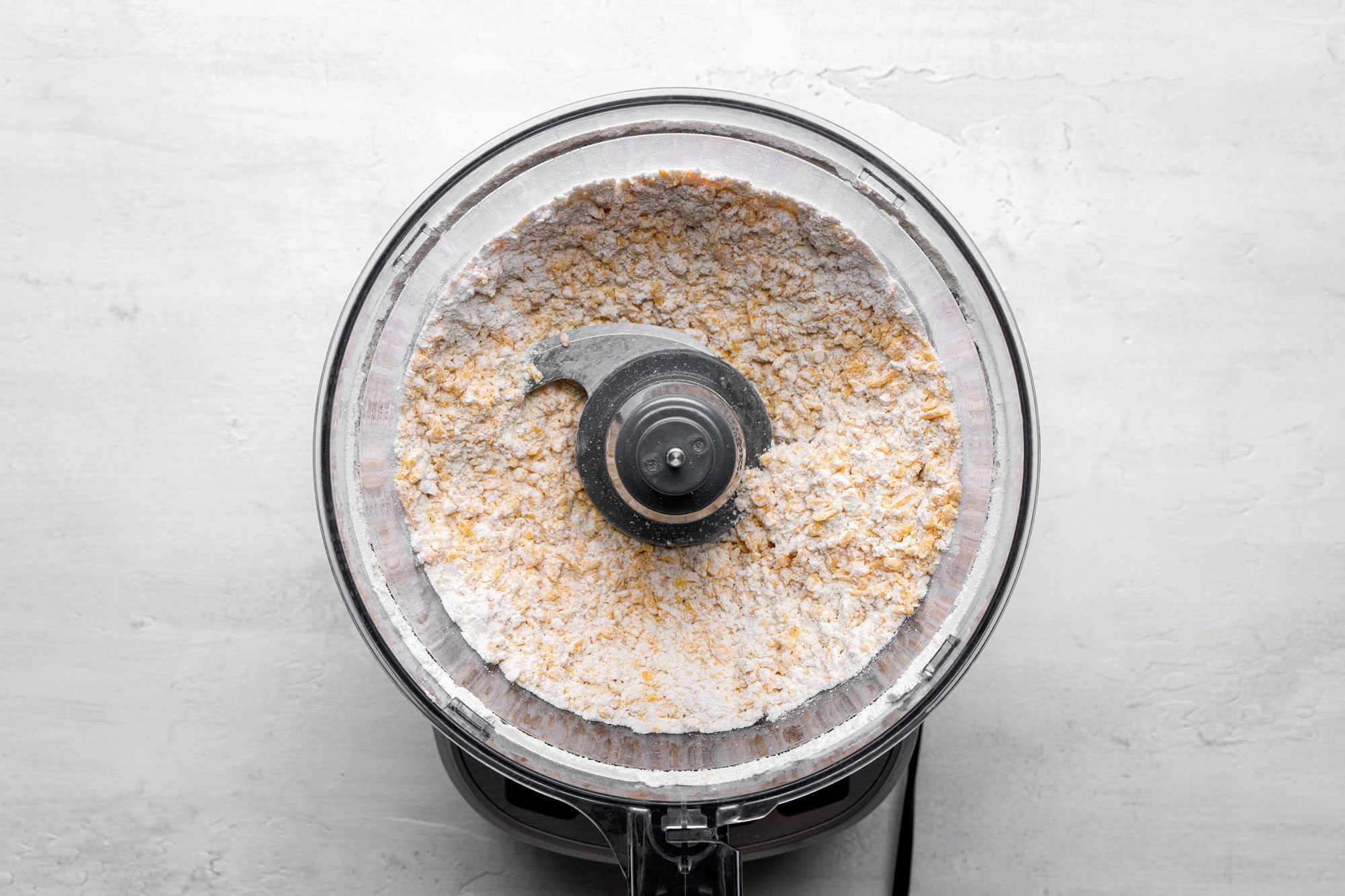 A top shot of a food processor contains a crumbly mixture of flour and butter, photographed from above on a light gray surface; this scene signifies the initial stage of dough preparation in baking