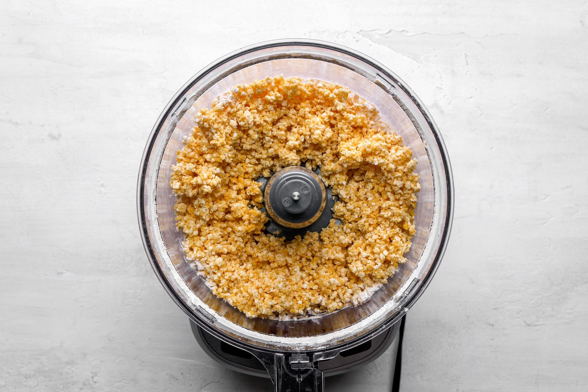 A top shot of the food processor; containing finely chopped cauliflower resembling rice; sits on a light-colored countertop. The background is neutral and uncluttered; highlighting the fresh cauliflower preparation