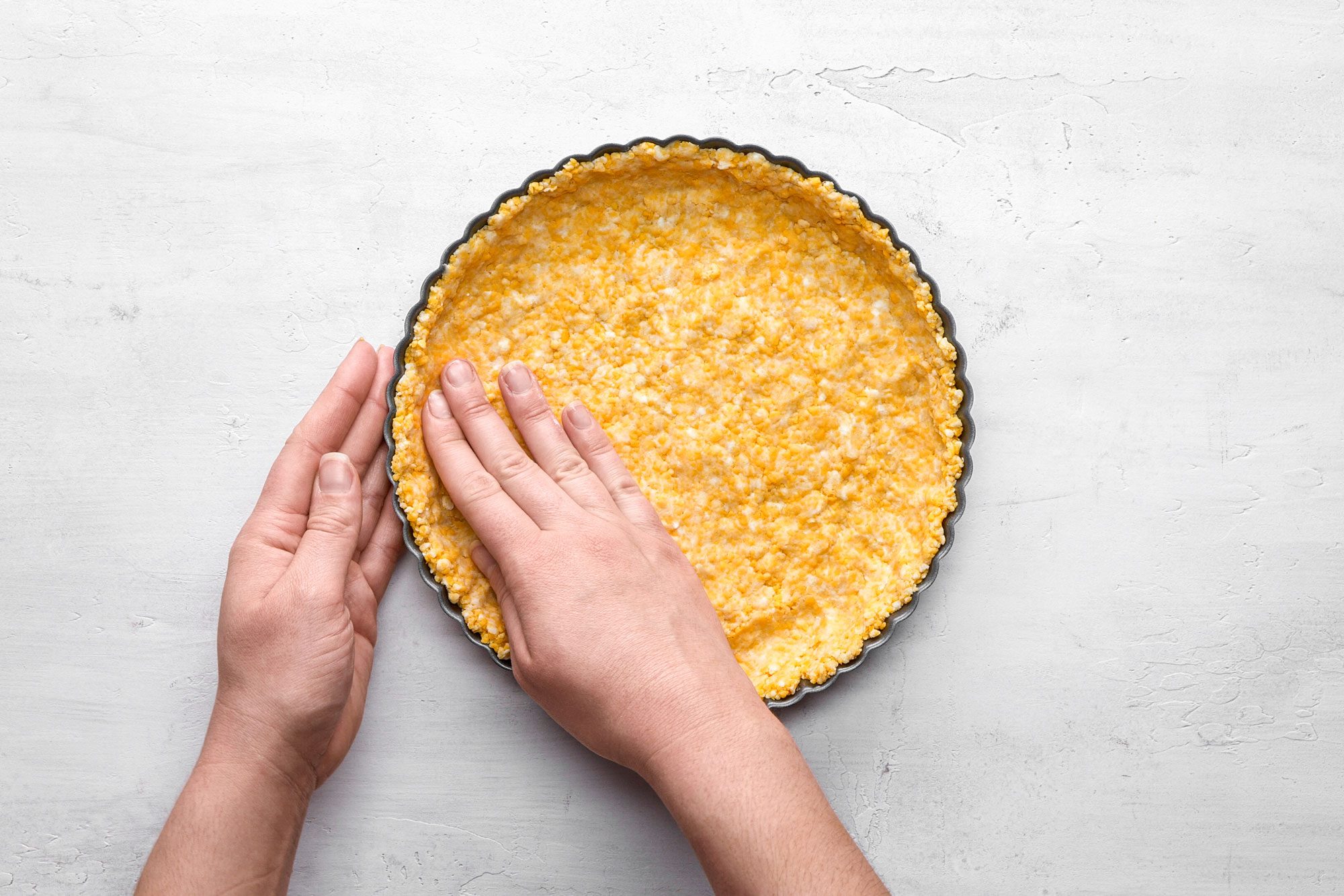 A top shot of Two hands evenly press dough into a round tart pan; on a light gray background; preparing crust for baking
