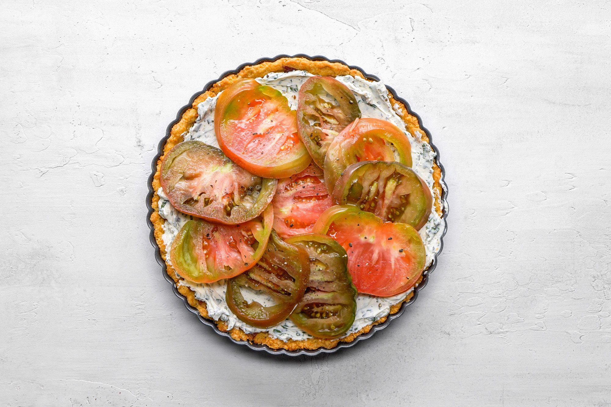 A Shot of a vibrant tart features overlapping red and green heirloom tomato slices; the colorful arrangement sits atop a white textured background, highlighting freshness and rustic appeal