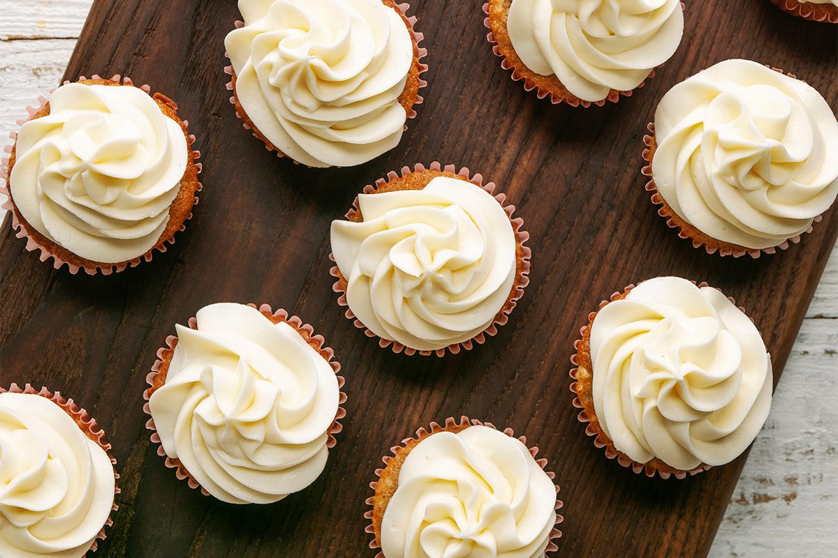 Nine cupcakes with swirled white frosting are arranged on a dark wooden surface, seen from above. The cupcakes are spaced out, with some on the board and others partially off to the side.
