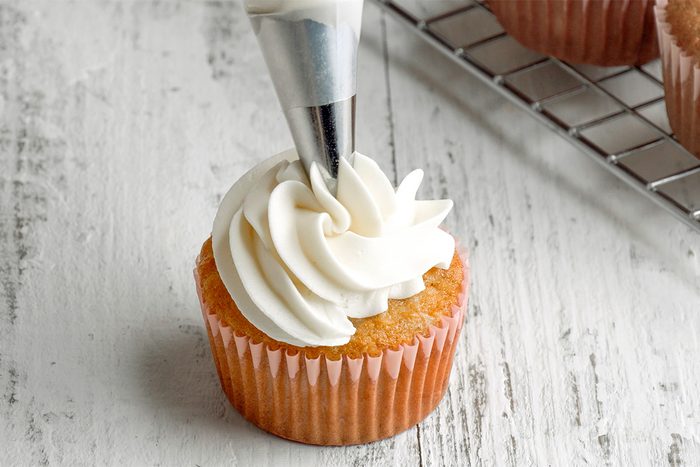 A vanilla cupcake is being decorated with white frosting using a piping bag on a white wooden surface, with more cupcakes on a cooling rack in the background.