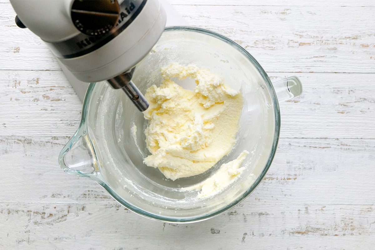 A stand mixer bowl with creamed butter and sugar, viewed from above on a white wooden surface. The mixer attachment is partially visible on the left side of the bowl.