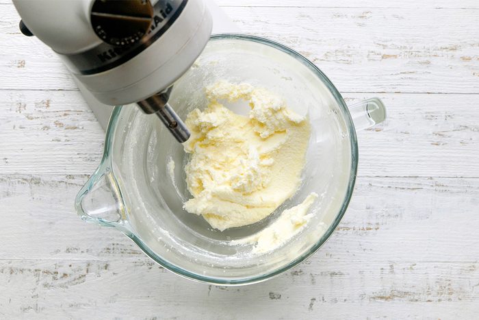 A stand mixer bowl with creamed butter and sugar, viewed from above on a white wooden surface. The mixer attachment is partially visible on the left side of the bowl.