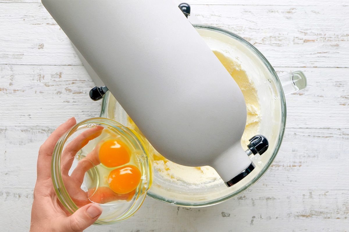 A hand holds a small glass bowl with two raw eggs above a stand mixer containing a light batter, ready to add the eggs. The scene is set on a white wooden surface.