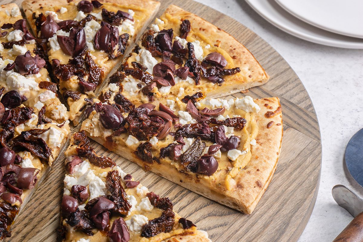 A sliced Mediterranean-style pizza topped with olives, sun-dried tomatoes, and crumbled cheese sits on a wooden platter next to stacked white plates and a pizza server.