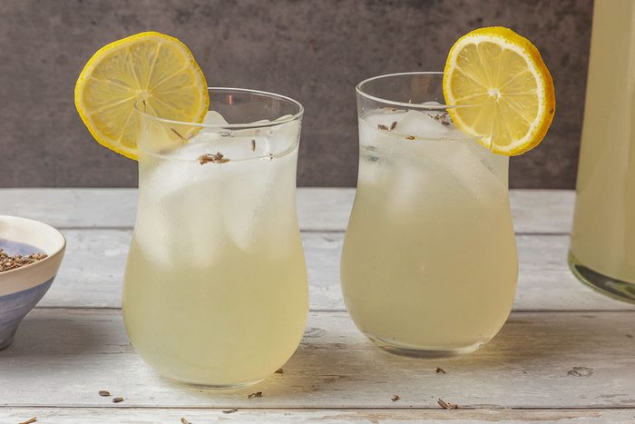 Front shot of Two lemonade glasses with ice and lemon slices rest on a light wooden surface; a pitcher and bowl of dried herbs appear blurred behind; the scene feels fresh and summery