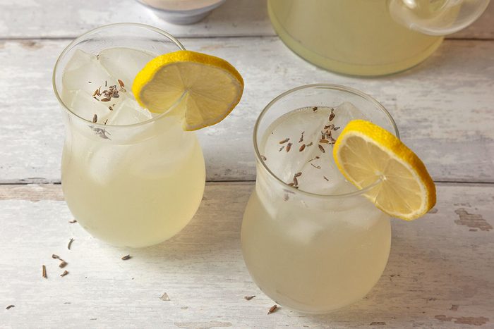 3/4th Shot of a Two glasses of lemonade with ice; lemon slices and lavender buds garnish them; beside a pitcher on a rustic white wooden surface