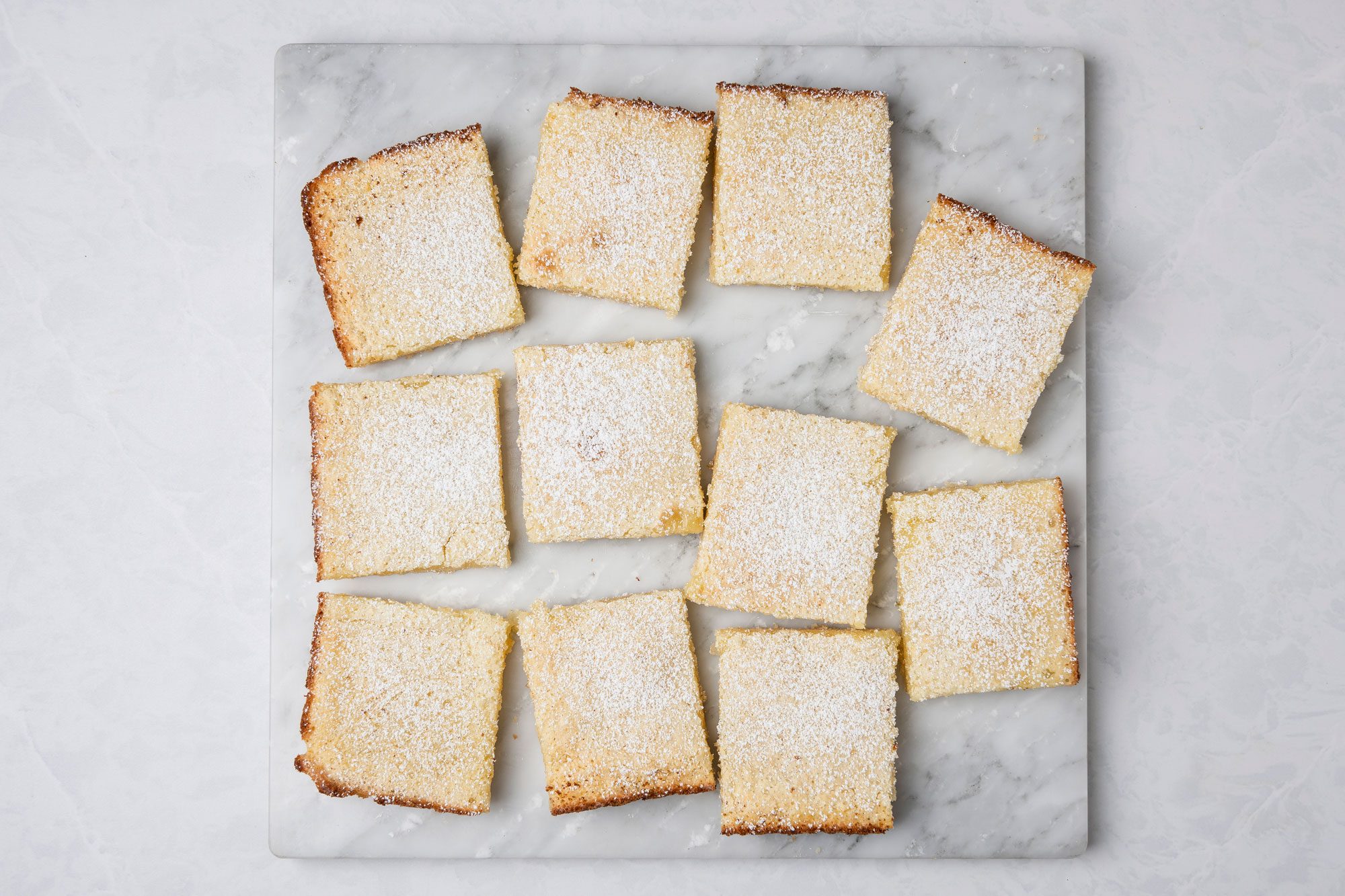 A top shot of a Nine square lemon bars dusted with powdered sugar sit in a grid on a white marble surface; their golden-brown edges and neat spacing highlight their homemade appeal and freshness