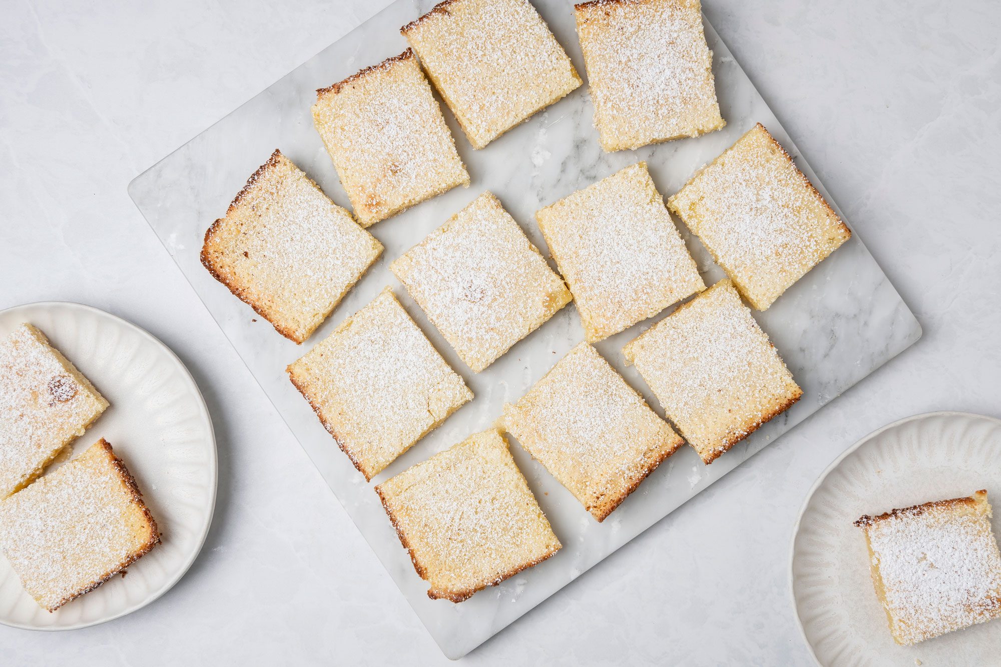 Top shot of Two lemon bars dusted with powdered sugar rest on a white plate; additional bars and a fork appear blurred in the background; bright lighting enhances the dessert's yellow color