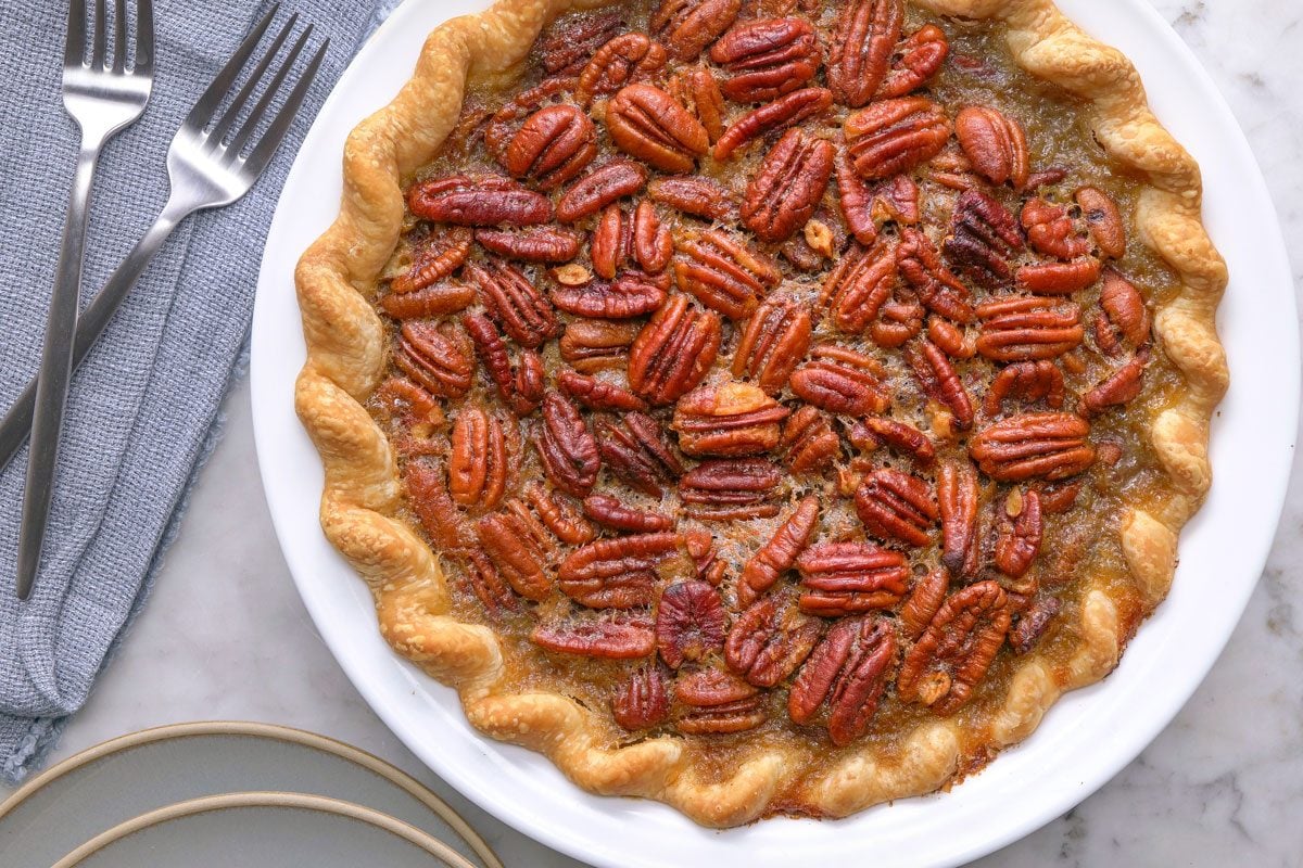 overhead shot of a whole pecan pie with a golden brown crust sits on a white plate, Three forks rest on a folded gray napkin nearby, and two stacked plates are partially visible on a marble surface