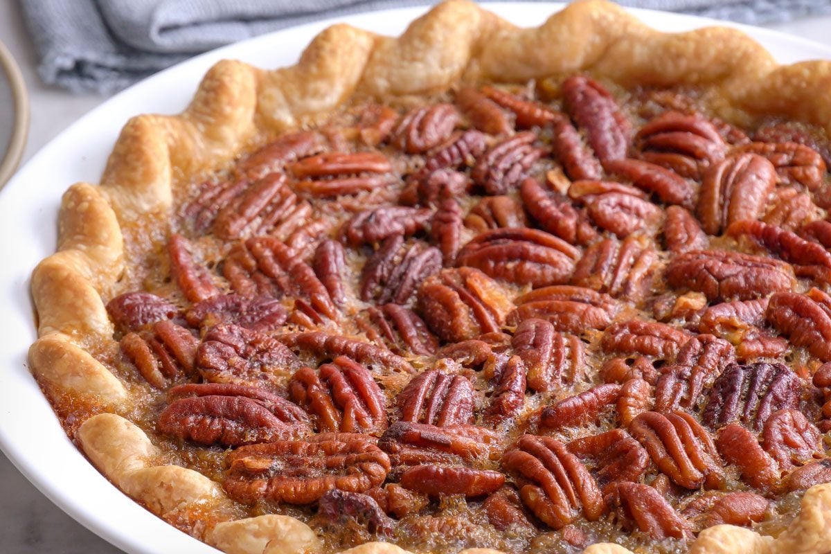 closeup shot of a baked pecan pie in a white pie dish, featuring a golden, crimped crust and a glossy layer of whole pecans on top