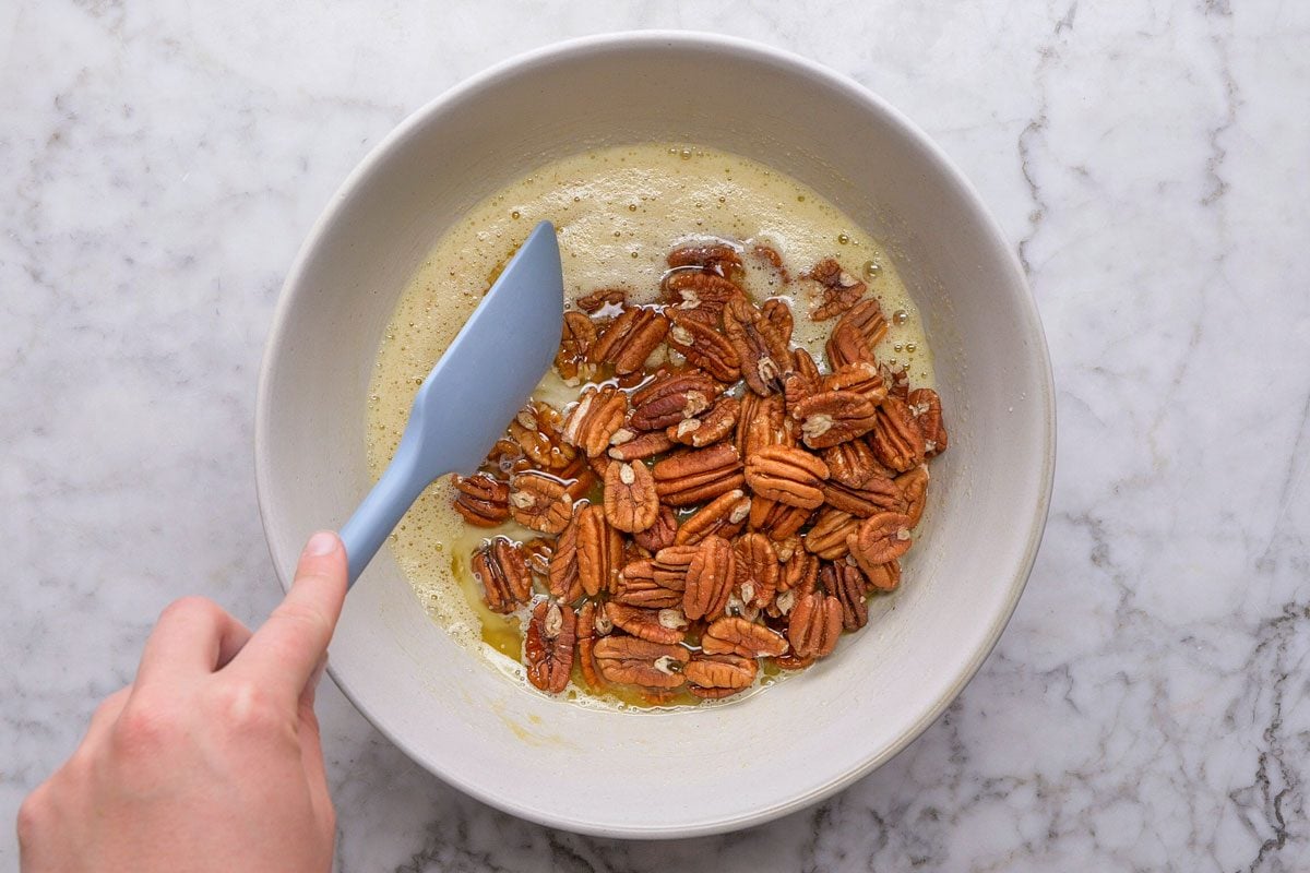 overhead shot of a hand stirs whole pecans into a bowl of frothy liquid with a light blue spatula, on a white marble countertop