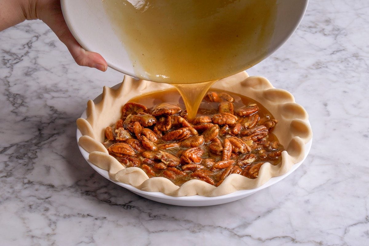 3/4th shot of a hand pours a golden brown liquid mixture over pecans in an unbaked pie crust, preparing a pecan pie on a marble countertop