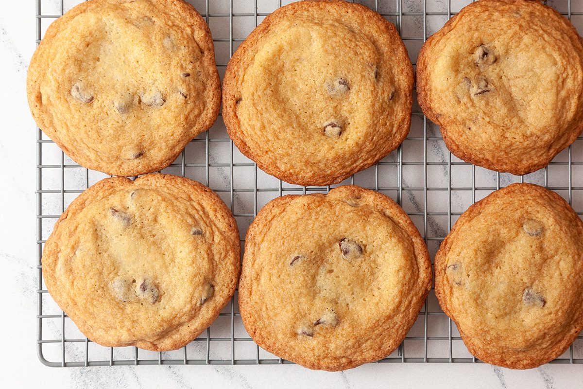 Six golden brown chocolate chip cookies cooling on a wire rack, viewed from above on a light-colored surface.