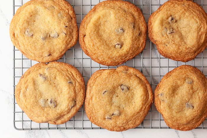 Six golden brown chocolate chip cookies cooling on a wire rack, viewed from above on a light-colored surface.