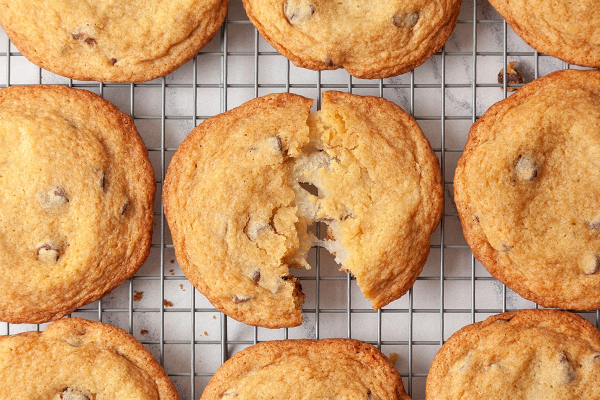 A batch of golden-brown chocolate chip cookies cooling on a wire rack, with one cookie in the center broken in half to reveal its soft interior.