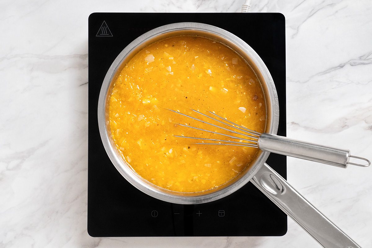 A saucepan filled with a yellow liquid mixture is on a black stovetop, with a metal whisk resting inside, all set on a white marble countertop.