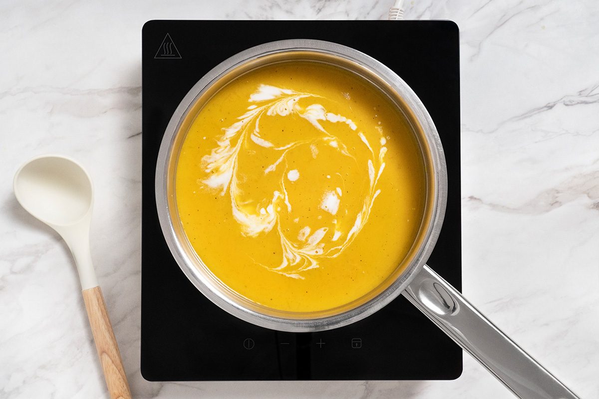 A saucepan filled with orange soup topped with a swirl of cream sits on an electric stovetop, next to a white spoon with a wooden handle on a marble countertop.