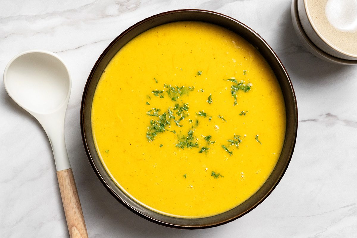 A bowl of creamy yellow soup garnished with chopped herbs sits on a marble surface, next to a white spoon with a wooden handle and a small bowl.